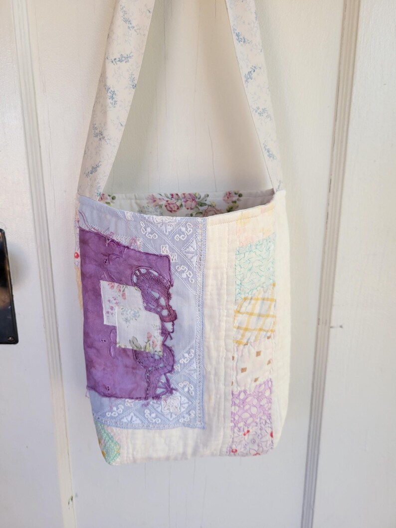May include: A white patchwork tote bag with a purple and blue floral print. The bag has a long strap and is made of various fabrics.
