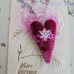 May include: A heart-shaped pendant necklace with a burgundy velvet texture, adorned with a floral embellishment. The necklace features a silver chain and small pink beads. The pendant is set on a vintage postcard with the words "Happy Returns".