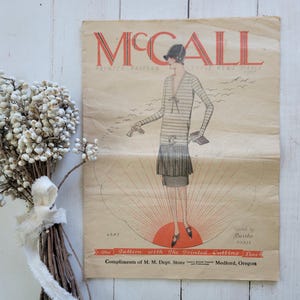 May include: Vintage McCall's printed pattern magazine cover. The cover features a fashion illustration of a woman in a striped dress. The magazine is on a white wooden surface, with a bouquet of white flowers tied with a ribbon.