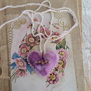 May include: A purple velvet heart pendant necklace with a gold star accent. The necklace has a white and pink beaded chain. The pendant rests on a vintage floral card with the words "Best Regards".