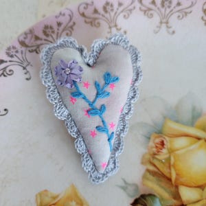 Puede incluir: Broche de tela en forma de corazón con borde de ganchillo gris. El broche presenta bordados de vides azules, estrellas rosas y una flor morada. El broche está sobre un plato decorativo con un diseño floral.