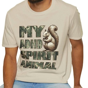 Puede incluir: Una camiseta beige con un estampado gráfico de una ardilla y el texto "MY ADHD SPIRIT ANIMAL". El texto está en un patrón de camuflaje.