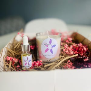 May include: A white gift box filled with shredded paper, a small bottle of essential oil, and a candle with a pink and purple label that says "Serenity Essence" and "Beauty Sleep for Soul".
