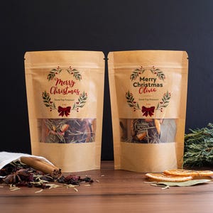 May include: Two brown paper pouches with clear windows, filled with potpourri. The pouches have a "Merry Christmas" design with a wreath and bow. The text "Stove Top Potpourri" is also visible. The pouches are on a wooden surface.