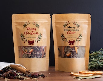 May include: Two brown paper pouches with clear windows, filled with potpourri. The pouches have a "Merry Christmas" design with a wreath and bow. The text "Stove Top Potpourri" is also visible. The pouches are on a wooden surface.