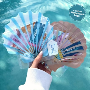 May include: Two shell-shaped pouches, one light blue and one tan, each featuring a clear window revealing various small packets inside. A white tag attached to the blue pouch displays the text "I got ship-faced" with small blue icons.