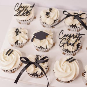 May include: A dozen cupcakes with white frosting and black decorations, including bows, sprinkles, and graduation-themed toppers. Some cupcakes have the text "Class of 2026" and "Congrats Grad".