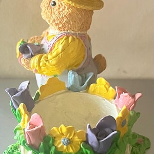 May include: A decorative Easter bunny figurine wearing a yellow hat and holding a basket. The bunny is surrounded by a floral arrangement with pink, yellow, and purple flowers, and a white picket fence. The base is green.