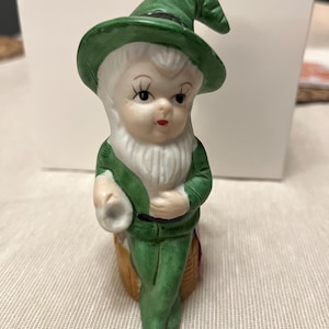 May include: A ceramic figurine of a leprechaun. The leprechaun has a green hat and outfit, a white beard, and a basket. The figurine is painted with details like eyelashes and a red mouth. The figurine is a decorative item.