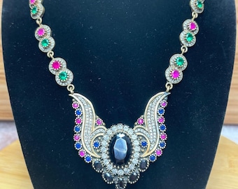 May include: A silver necklace with a large black gemstone pendant surrounded by smaller white, blue, and pink gemstones. The necklace has a chain with alternating green and pink gemstones.
