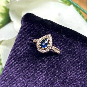 May include: A gold-toned ring with a pear-shaped, deep blue gemstone at its center. The gemstone is surrounded by a halo of smaller, clear stones. The ring band features delicate filigree detailing, creating an elegant and sophisticated look.
