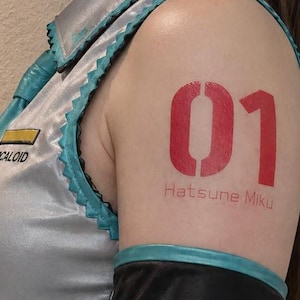 May include: A person's arm with a large red stencil tattoo of the number "01" and the text "Hatsune Miku". The person is wearing a silver and teal costume with the word "VOCALOID" visible.