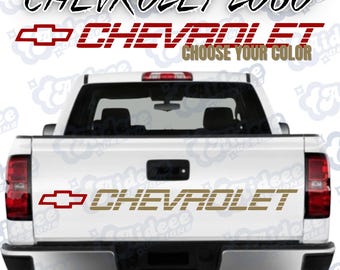 May include: A white pickup truck tailgate featuring a gold Chevrolet logo and text decal. The text reads CHEVROLET LOGO, CHEVROLET, CHOOSE YOUR COLOR, and CHEVROLET. The design includes the iconic bowtie emblem in red and gold, applied to the center of the tailgate.