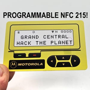 May include: A rectangular yellow programmable NFC 215 card featuring a graphic design of a vintage mobile phone interface. The screen displays the text GRAND CENTRAL HACK THE PLANET. The Motorola logo and directional navigation buttons are printed below.