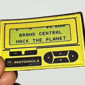 May include: A rectangular yellow embroidered patch featuring a digital pager design. The screen displays the text GRAND CENTRAL HACK THE PLANET in black. Below the screen, the Motorola logo and navigation buttons are depicted in black thread.