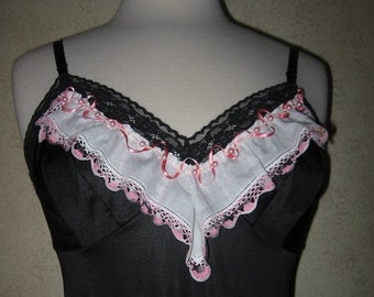May include: Black slip dress with a pink ribbon belt and a decorative black button. The bodice features a white and pink ruffled trim with black lace and pink ribbon accents. The dress is displayed on a mannequin.