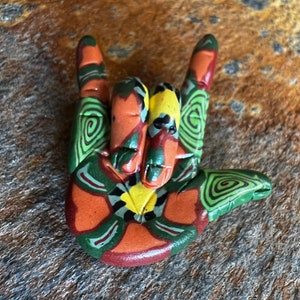 Könnte beinhalten: Eine handgefertigte Tonskulptur einer Hand, die das amerikanische Zeichensprachzeichen für "Ich liebe dich" macht. Die Skulptur ist in einem farbenfrohen Muster aus Grün, Orange, Gelb und Schwarz bemalt.