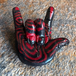 Könnte beinhalten: Eine schwarze und rote Handskulptur in Form des "Ich liebe dich"-Handzeichens. Die Hand ist mit schwarzen und roten Streifen bemalt.