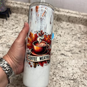 Op de afbeelding: Een wit en bruin geïsoleerde tumbler met een deksel en een rietje. De tumbler heeft een "Coffee Weather" ontwerp met een koffiekopje, herfstbladeren en koffiebonen. De tumbler wordt in een hand gehouden.