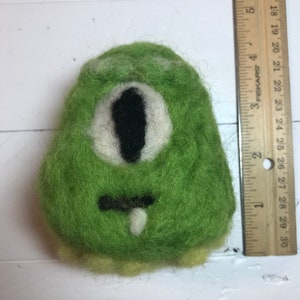 May include: A green felt creature with a single large eye and a small mouth. The creature is about 3 inches tall.