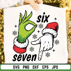 May include: White t-shirt with a graphic design featuring a green hand making an "okay" gesture and a white hand pointing down, both wearing red and white Santa cuffs. The words "six" and "seven" are above and below the hands, with snowflakes.
