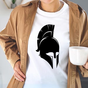 May include: A white t-shirt with a black silhouette of a Spartan helmet. The helmet has a tall, pointed crest and a face mask with a prominent nose guard. The t-shirt is being worn by a person with a brown jacket.