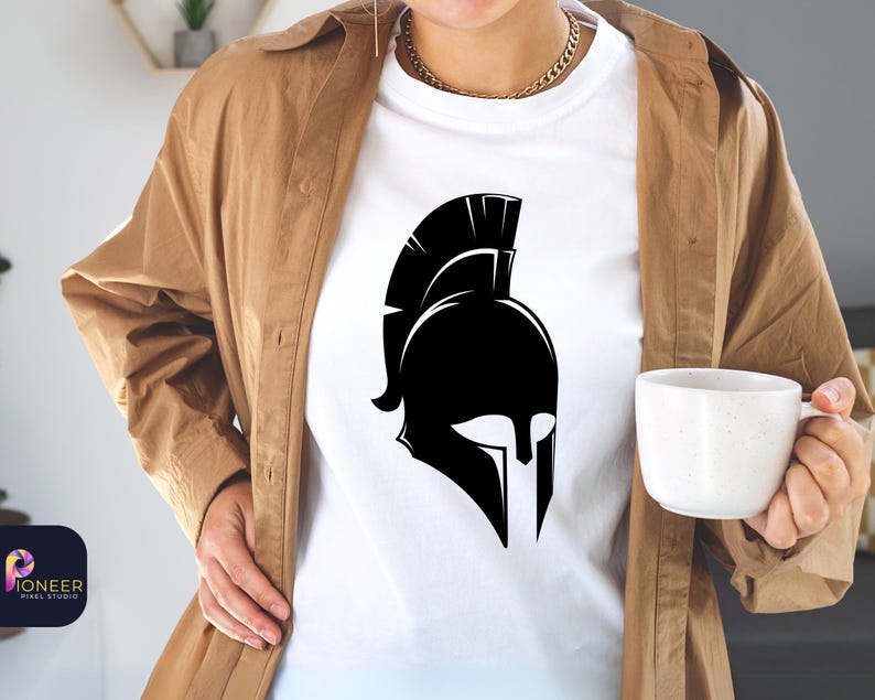 May include: A white t-shirt with a black silhouette of a Spartan helmet. The helmet has a tall, pointed crest and a face mask with a prominent nose guard. The t-shirt is being worn by a person with a brown jacket.