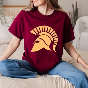 May include: A maroon t-shirt with a yellow graphic of a Spartan helmet. The helmet has a plume of feathers on top.