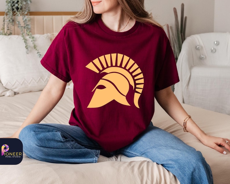 May include: A maroon t-shirt with a yellow graphic of a Spartan helmet. The helmet has a plume of feathers on top.