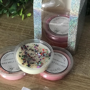 May include: Wax melt pots in clear plastic containers, some with glitter. The labels read "SCENTYM" and "WAX MELT DELI". A box of melts is visible in the background. The melts are for use in a wax melt burner.