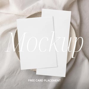 Könnte beinhalten: Zwei leere weiße Karten auf einem weißen Keramikteller mit weißem Stoffhintergrund. Der Text "Mockup" ist in Weiß auf dem Bild geschrieben. Der Text "FREE CARD PLACEMENT" ist in Schwarz am unteren Rand des Bildes geschrieben.