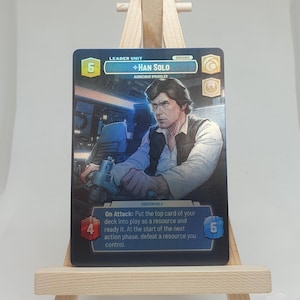 May include: A blue and silver Star Wars trading card featuring Han Solo, a character from the Star Wars franchise. The card has a picture of Han Solo in the Millennium Falcon cockpit. The card text reads "Leader Unit + Han Solo Audacious Smuggler Ground Underworld On Attack: Put the top card of your deck into play as a resource and ready it. At the start of the next action phase, defeat a resource you control."