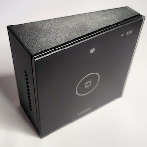 Puede incluir: Un timbre negro con un botón blanco y una pequeña lente de cámara. El timbre tiene una superficie texturizada y una pequeña rejilla de altavoz en el lateral.