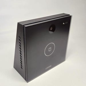 Può includere: Un campanello rettangolare nero con una fotocamera, un'icona a forma di campana e il marchio "LOXONE" stampato sulla parte anteriore. Il dispositivo ha un design elegante e moderno con finitura lucida e cornice nera opaca. Il campanello è montato su una superficie bianca.