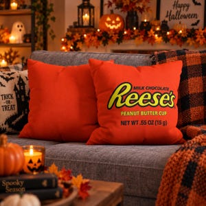 May include: A square orange throw pillow featuring the Reese's Milk Chocolate Peanut Butter Cup logo in yellow and brown text, including the printed text NET WT .55 OZ (15 g). The pillow is positioned on a gray sofa next to a black and white pillow.