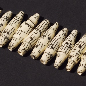 Könnte beinhalten: Nahaufnahme einer Perlenkette aus aufgerollten Musikblättern. Die Perlen sind alle unterschiedlich lang und haben eine cremefarbene Farbe mit schwarzen Noten darauf gedruckt.