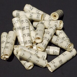 May include: A collection of small, white, rolled paper tubes with black Japanese characters printed on them. The tubes are arranged in a pile on a dark surface.