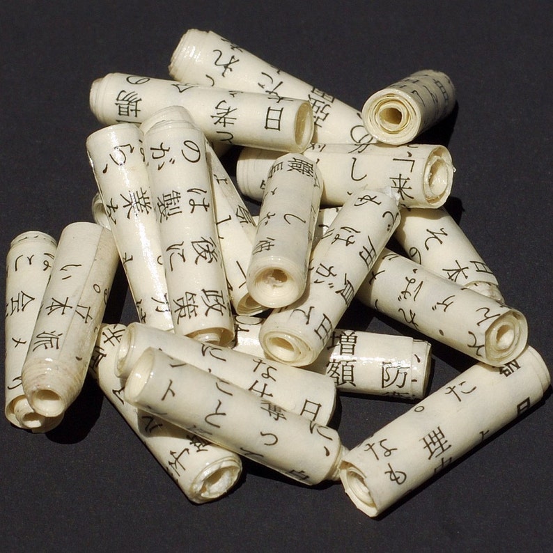 May include: A collection of small, white, rolled paper tubes with black Japanese characters printed on them. The tubes are arranged in a pile on a dark surface.