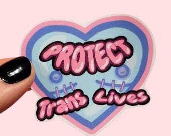 Può includere: Un adesivo a forma di cuore con un centro azzurro, un contorno blu scuro e un bordo rosa. Il testo "PROTECT" è scritto in lettere a bolla rosa nella parte superiore, e "Trans Lives" in basso, con punti di sutura rosa.