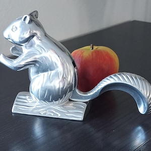 May include: A silver-colored squirrel nutcracker with a textured tail and a wooden base. A red and yellow apple rests beside the nutcracker on a dark surface. The squirrel is in a standing position.