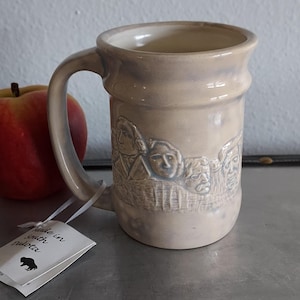 May include: A beige ceramic mug featuring a relief of Mount Rushmore. The mug has a large handle and a slightly flared rim. A tag attached to the handle reads "Made in South Dakota". An apple rests beside the mug.