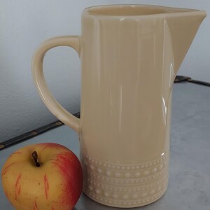 May include: A beige ceramic pitcher with a white embossed design around the bottom. The pitcher has a wide mouth and a handle.