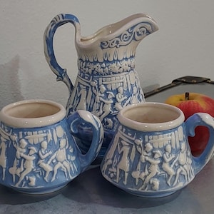 May include: A set of three blue and white ceramic cups and a pitcher with a whimsical design of people and houses. The cups have handles and the pitcher has a spout.
