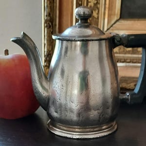 May include: A silver teapot with a black plastic handle. The teapot has a rounded body and a spout on the side. The teapot is sitting on a dark surface.