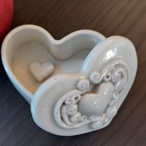 May include: White ceramic heart-shaped trinket box with a raised heart design and intricate details. The box is open and the inside is empty.