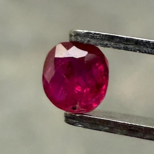May include: A faceted, oval-shaped ruby gemstone held by a pair of metal tweezers. The ruby is a deep, saturated red color with visible internal inclusions. The tweezers are silver-colored and provide a neutral background.