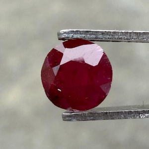 Peut inclure: Un rubis rond et facetté, maintenu par un outil métallique. Le rubis est d'un rouge profond avec des facettes visibles qui captent la lumière. La pierre précieuse semble polie et prête à être sertie dans un bijou.