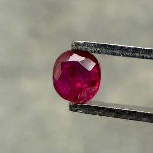 Op de afbeelding: Een gefacetteerde, ovaalvormige robijn edelsteen, vastgehouden door een pincet. De robijn heeft een diepe, levendige rode kleur met meerdere facetten die licht reflecteren. De pincet is zilverkleurig en houdt de edelsteen tegen een neutrale achtergrond.
