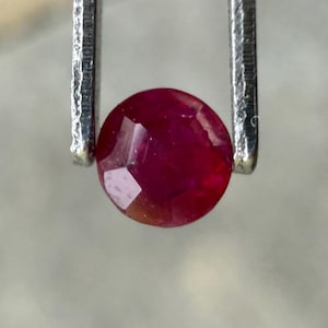 Op de afbeelding: Een gefacetteerde, ronde robijn edelsteen, vastgehouden door een zilveren pincet. De robijn heeft een diepe, rijke rode kleur met zichtbare interne facetten. De edelsteen is de focus van de afbeelding, die de helderheid en kleur laat zien.