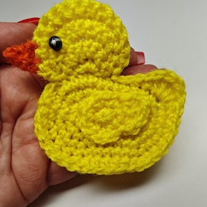 Könnte beinhalten: Eine handgefertigte gelbe Häkelente mit einem orangefarbenen Schnabel und einem schwarzen Auge. Die Ente ist eine kleine, dreidimensionale Figur, wahrscheinlich ein Spielzeug oder ein Dekorationsartikel. Die Häkelstiche sind sichtbar und verleihen ihr ein strukturiertes Aussehen.